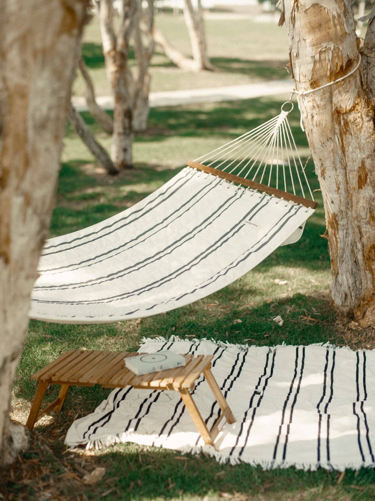 hammock hanging between two trees with a table and book underneath