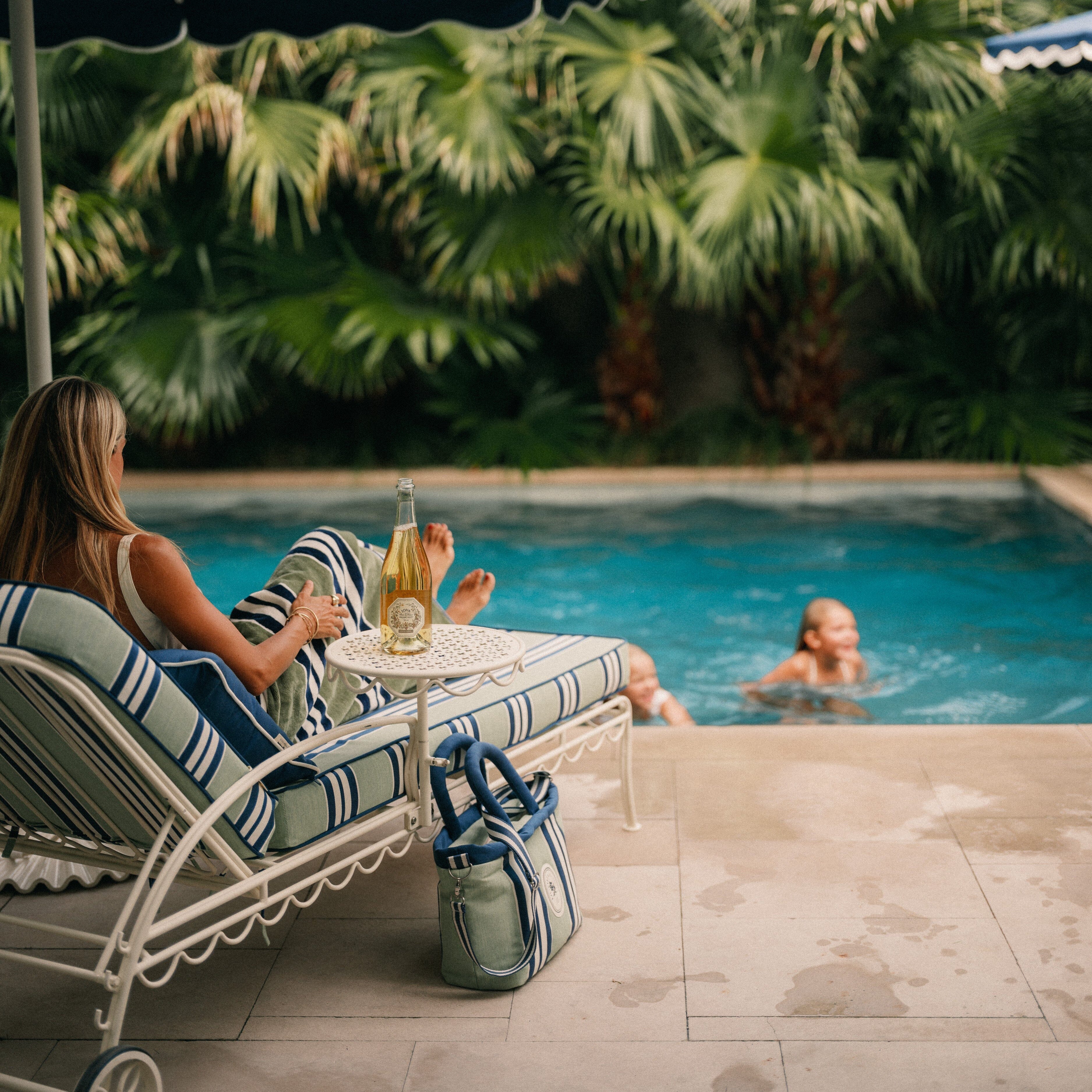 Woman sitting by a pool with a blue umbrella and another person in the water.