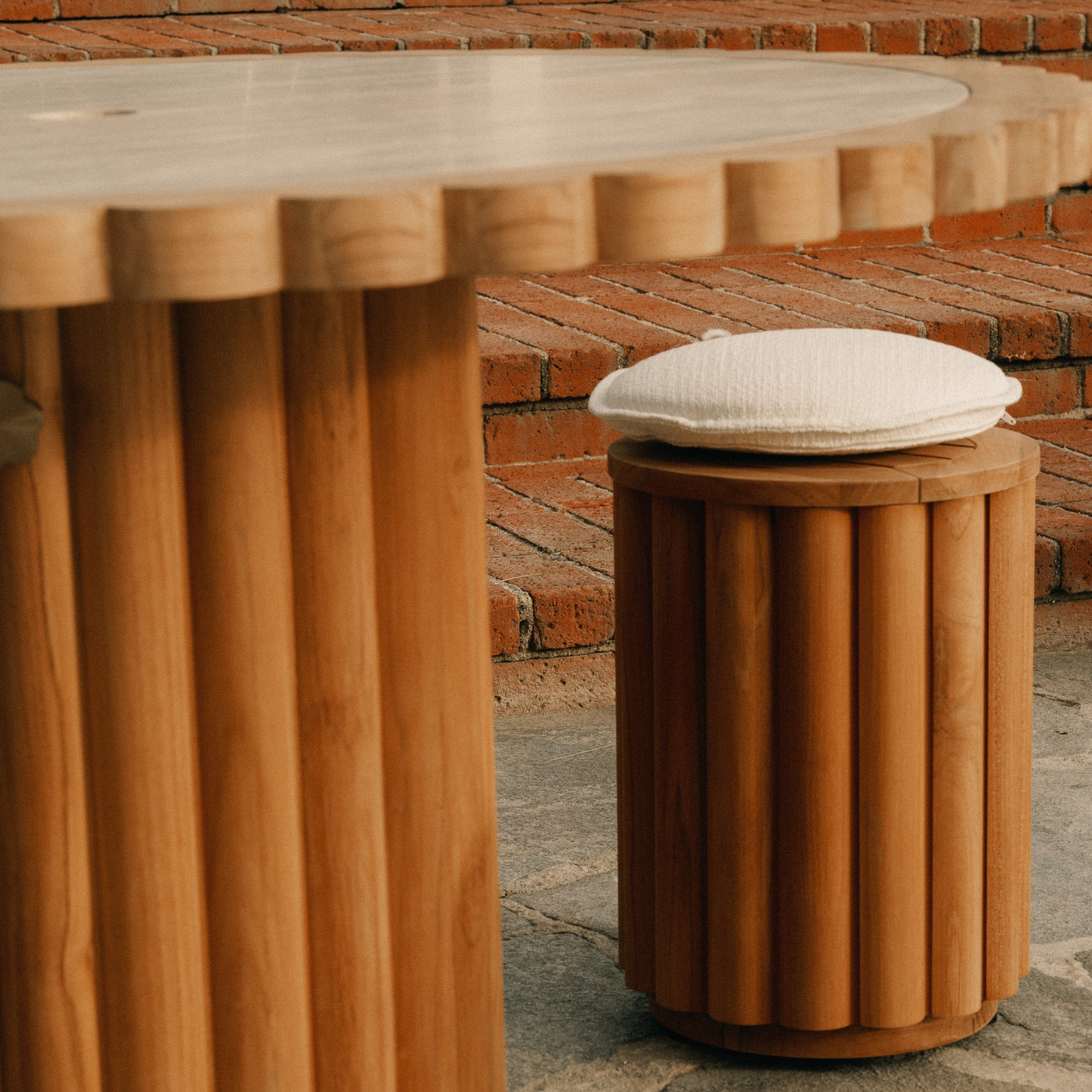 Wooden stool with a cushion next to a round wooden table on a stone surface with a brick wall background.
