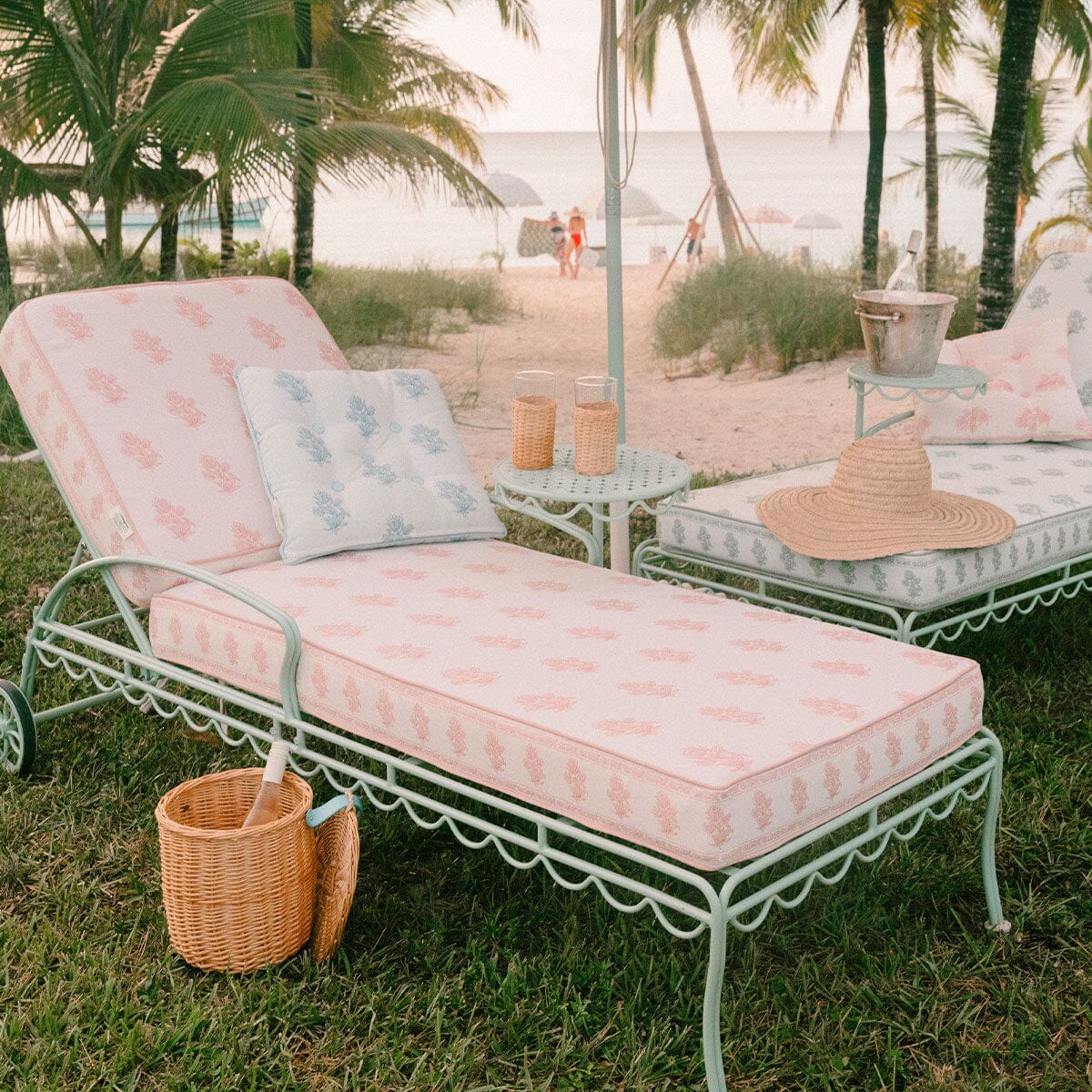 A pastel pink and white patterned metal beach daybed with a matching pillow is set on green grass at the beach, with palm trees, sand, and ocean in the background. A straw hat and a small wicker basket sit beside the daybed.