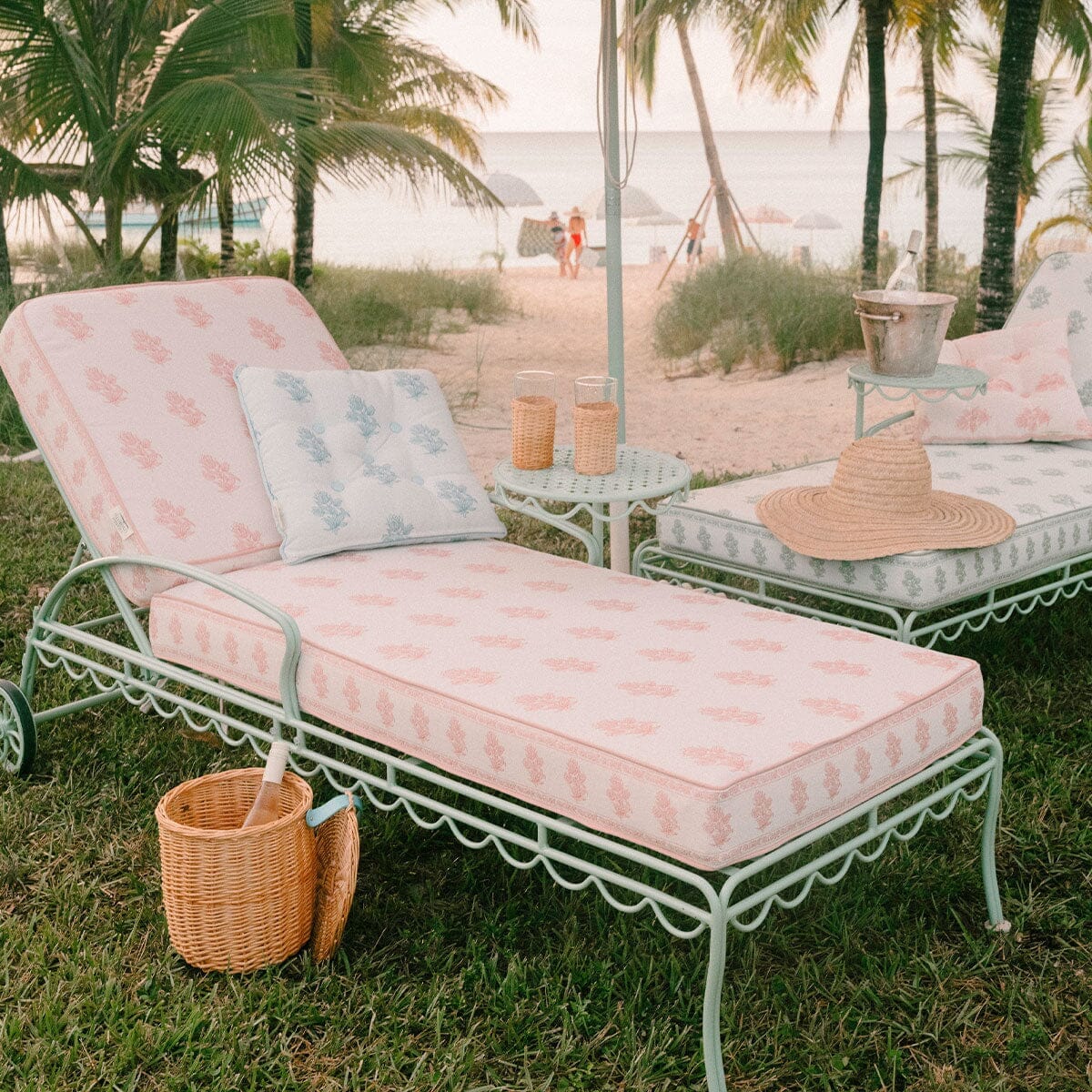 A pastel pink and white patterned metal beach daybed with a matching pillow is set on green grass at the beach, with palm trees, sand, and ocean in the background. A straw hat and a small wicker basket sit beside the daybed.
