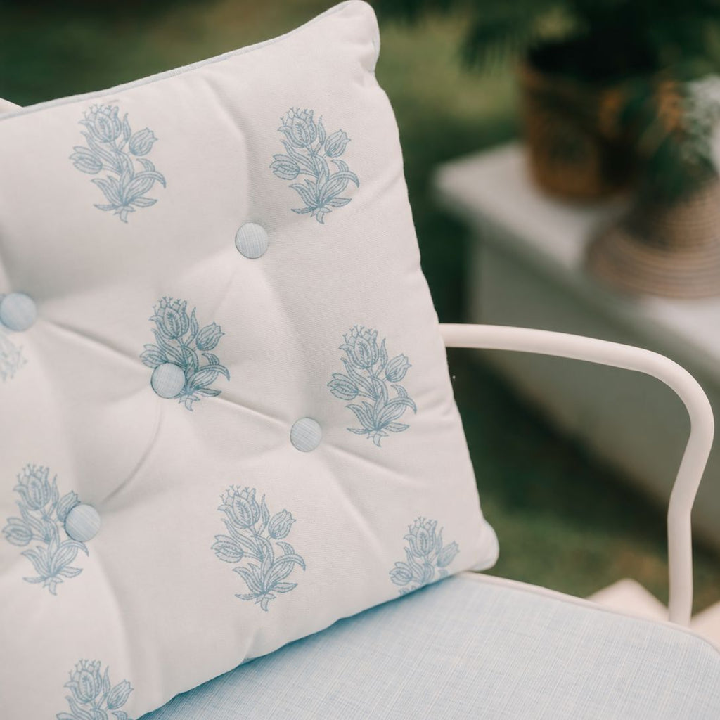 A close-up of a white tufted outdoor dining chair cushion with a light blue floral pattern, resting on a white metal chair in a garden setting.