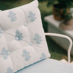 A close-up of a white tufted outdoor dining chair cushion with a light blue floral pattern, resting on a white metal chair in a garden setting.