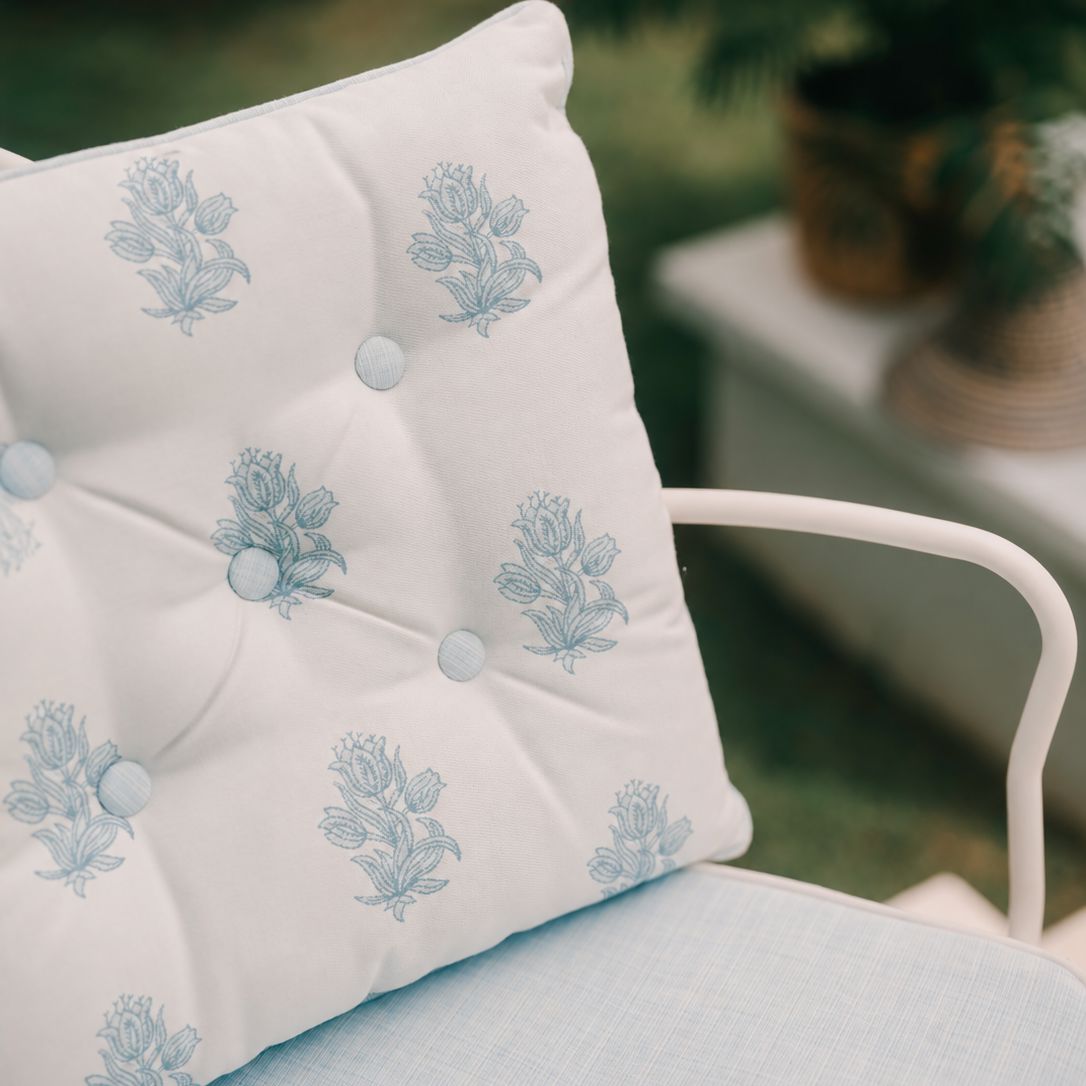 A close-up of a white tufted outdoor dining chair cushion with a light blue floral pattern, resting on a white metal chair in a garden setting.