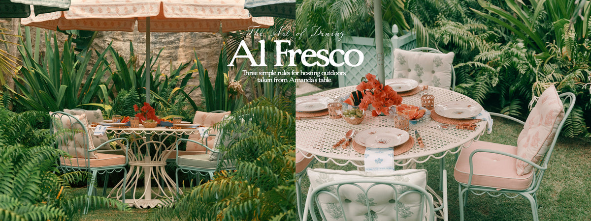Outdoor garden dining scene with a round white lace-top table, pink cushioned metal chairs, orange floral centerpieces, and tropical plants in the background. Large overlaid text reads 'Al Fresco' with smaller descriptive copy.