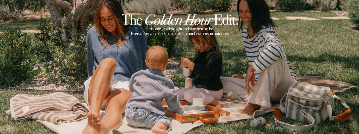 A family picnic scene outdoors on a grassy lawn, with two women and two children sitting on a striped blanket. A striped tote bag sits to the right, and the text 'The Golden Hour Edit' appears across the top with the tagline 'Cute little outfits and accessories to be Everything styled to make your first season summer fun.