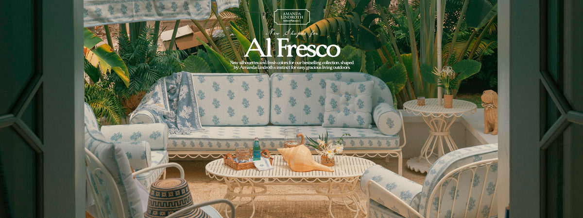 An outdoor patio scene viewed through open dark green doors, featuring a light blue and white floral patterned sofa, matching armchairs, a white wicker coffee table, and tropical green plants in the background. The text 'Al Fresco' is overlaid at the top center.