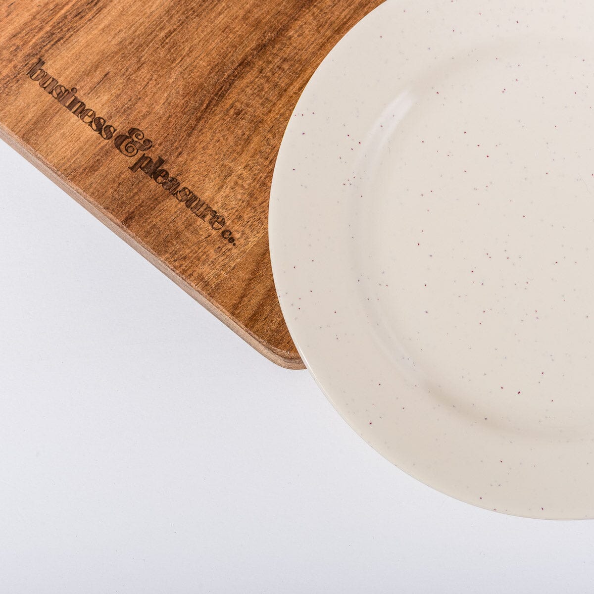 Wooden cutting board with 'pleasures & pleasures' engraving next to a ceramic plate on a white background