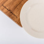 Wooden cutting board with 'pleasures & pleasures' engraving next to a ceramic plate on a white background