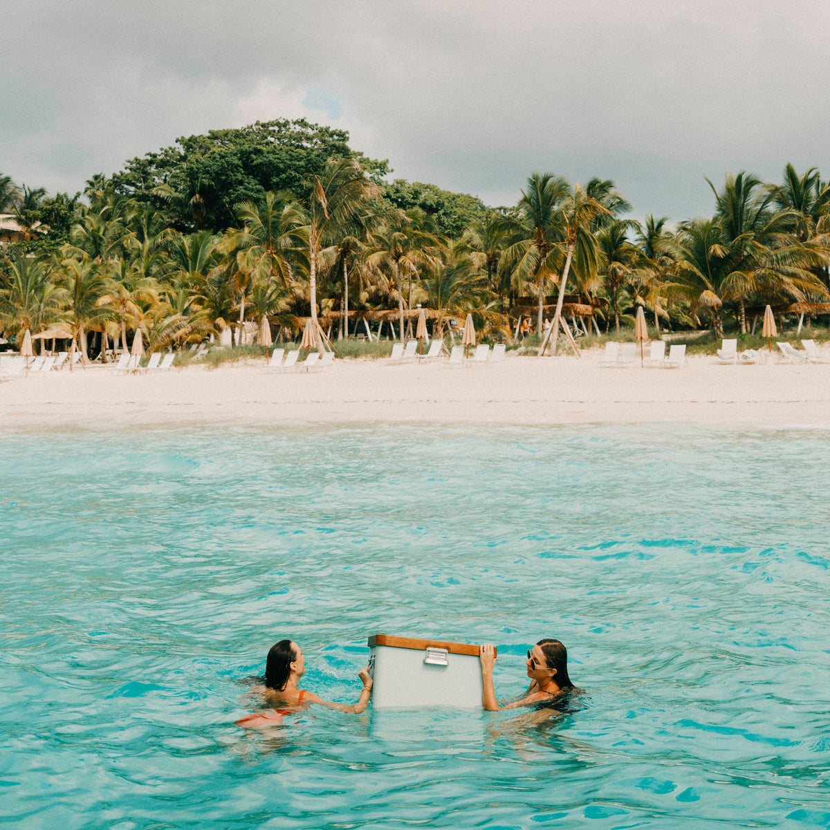 Two people swimming in turquoise ocean water holding a white rectangular board, with a sandy beach, palm trees, and a cloudy sky in the background.