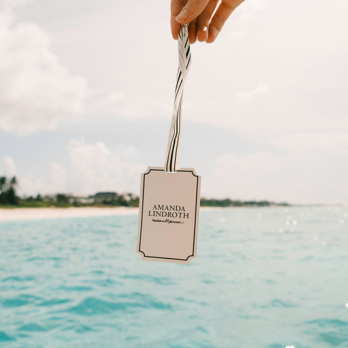 A hand holds a personalized wax melt tag with a silver twisted ribbon against a scenic background of turquoise ocean water and a cloudy sky. The tag is printed with the name 'AMANDA LINDROTH' 