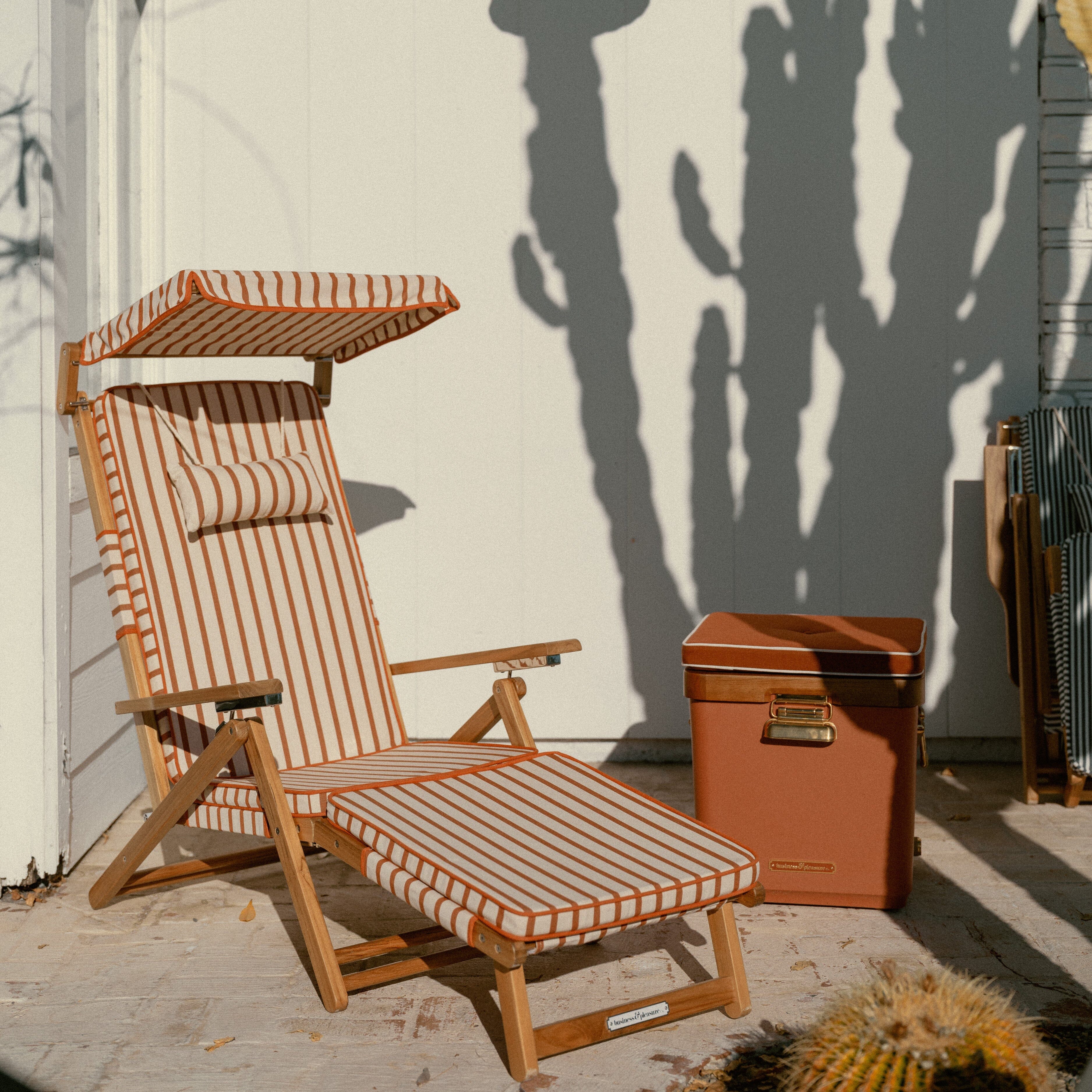Wooden lounge chair with striped cushion next to a cooler against a wall with cactus shadows.