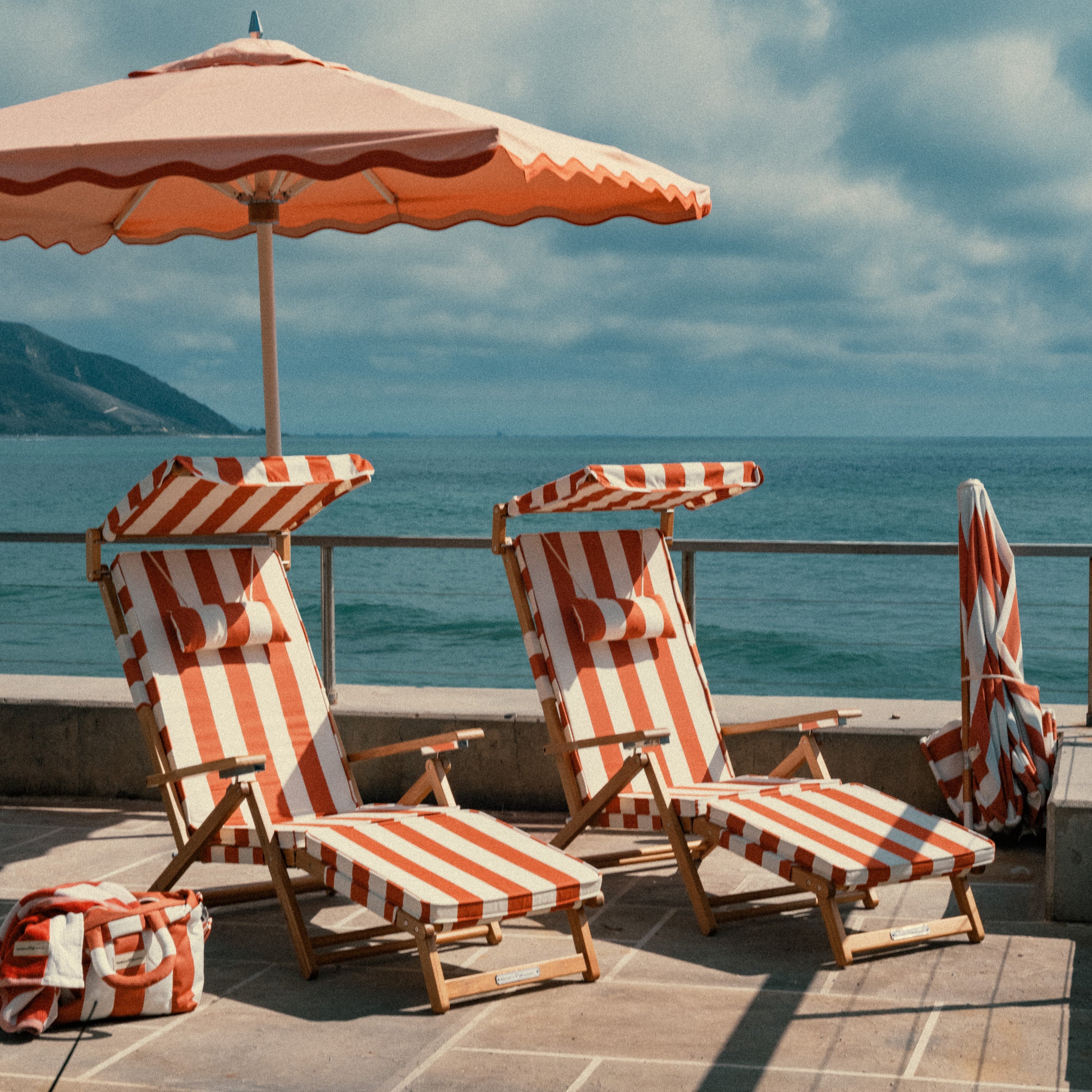 Beach chairs with striped covers and an umbrella on a deck overlooking the ocean.