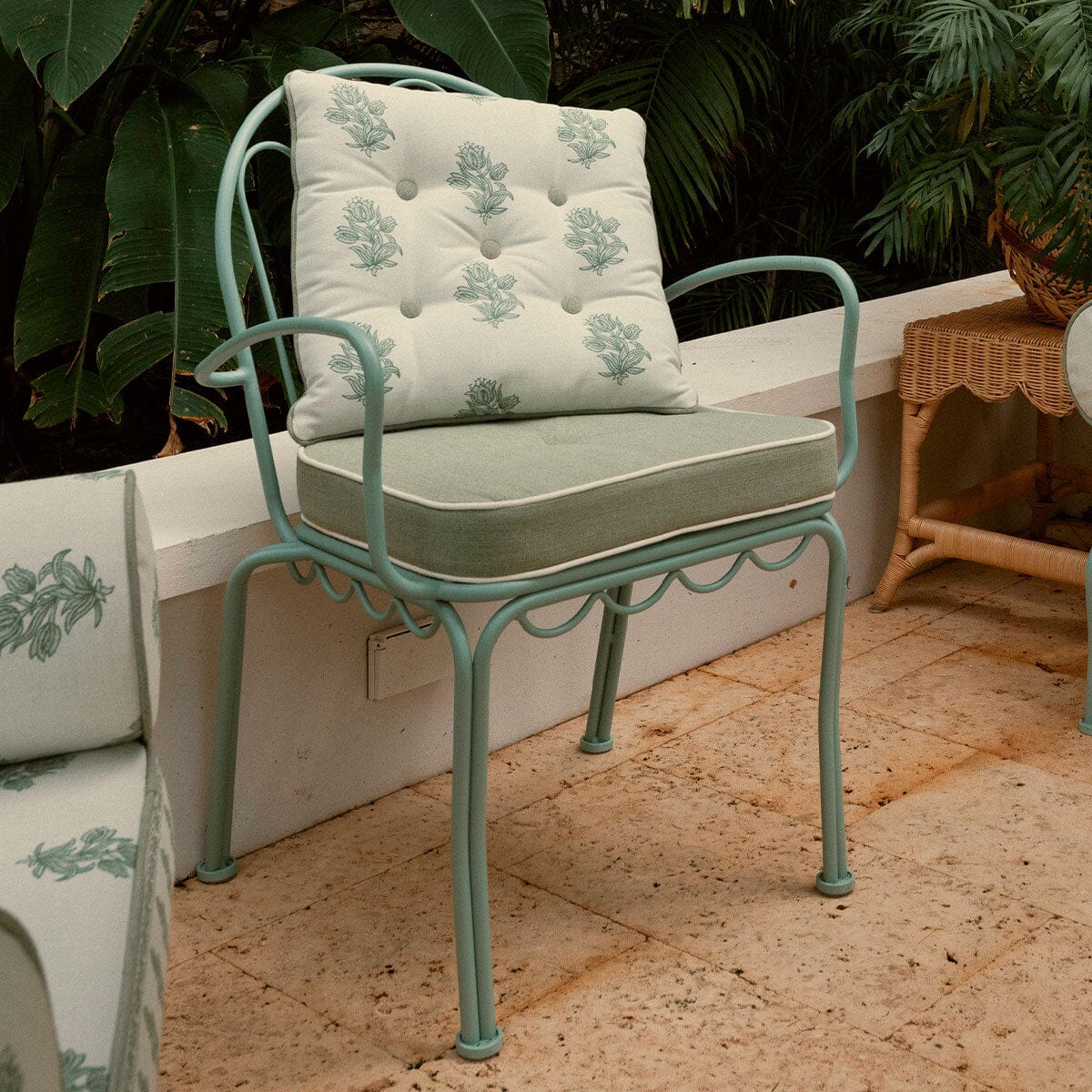 A mint green metal patio armchair with a tufted cream cushion featuring a sage green botanical pattern, set on a stone patio with tropical plants in the background.