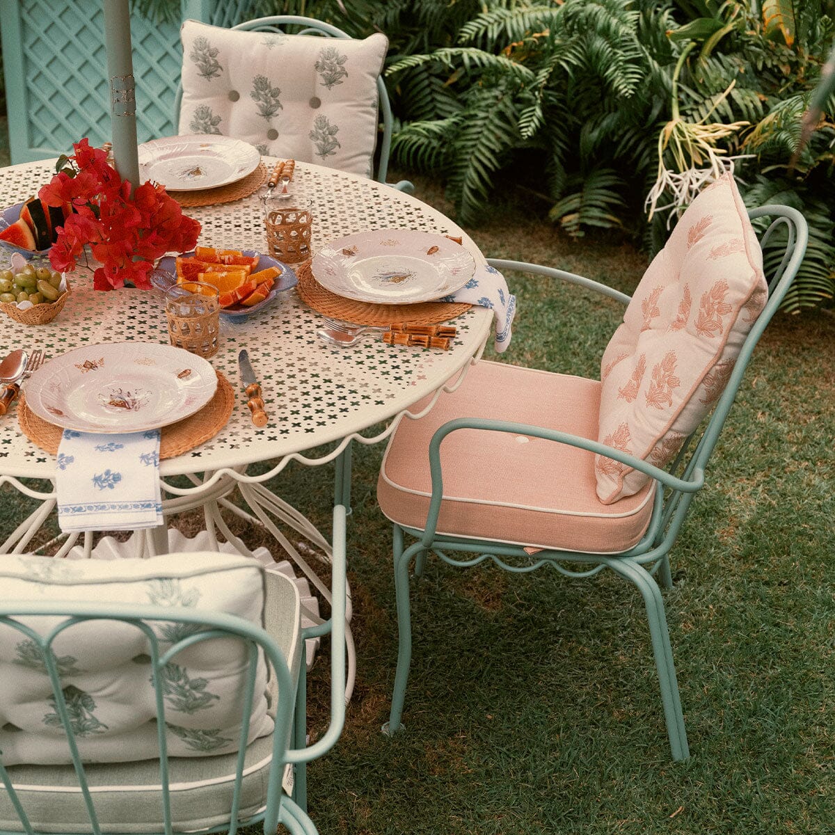 Outdoor patio dining set featuring a round white metal table and three matching light blue chairs with blush pink cushions, styled with plates, fruit, and red flowers on green grass with plants in the background.