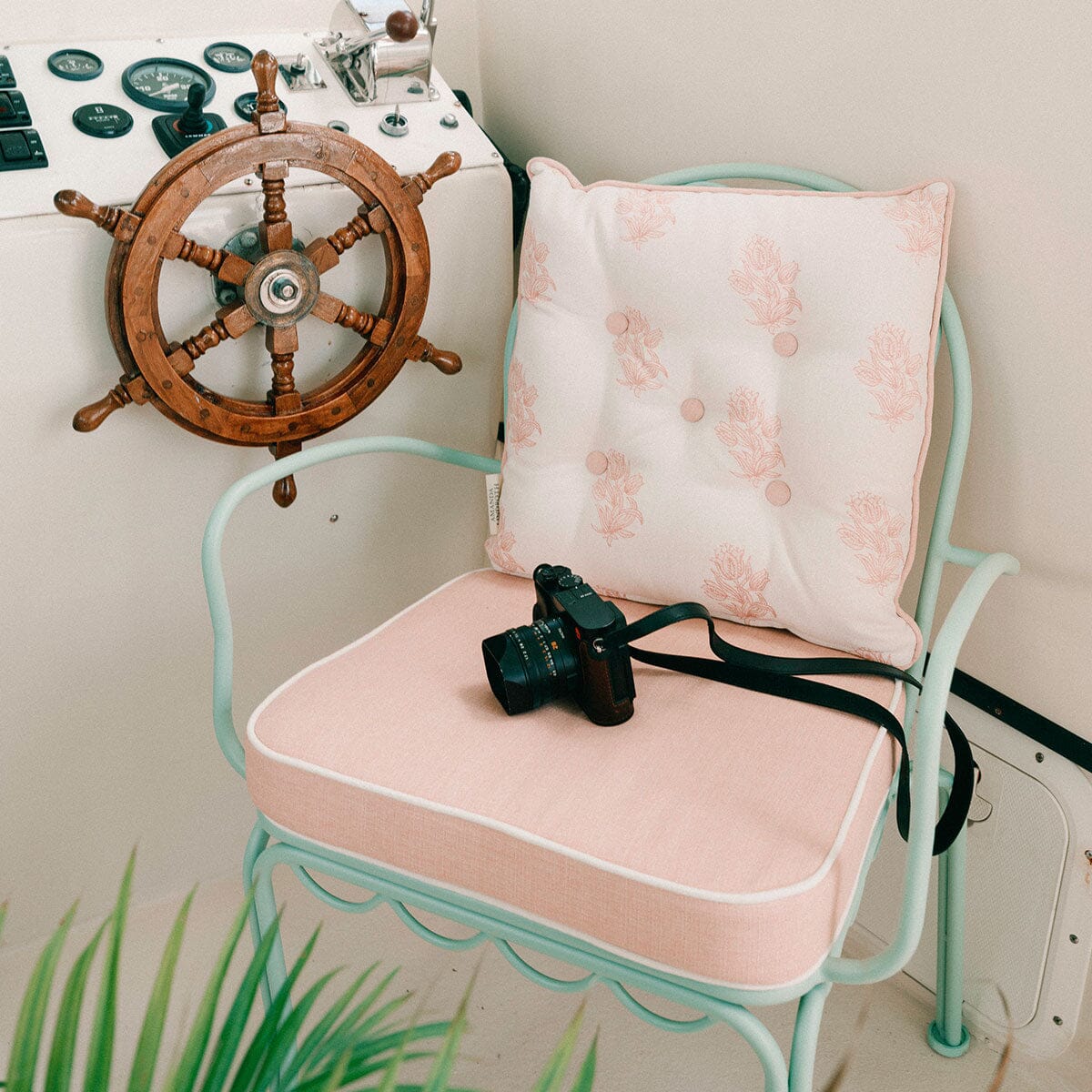 A mint green metal chair with two pale pink cushions, one on the seat and one on the backrest, styled with a wooden ship's wheel on the wall behind it and a black camera resting on the seat.