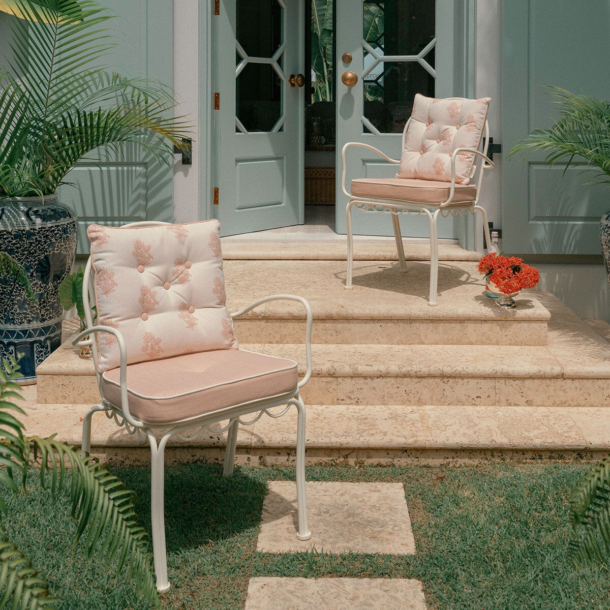 2metal patio armchairs with tufted cushions in a light beige and pink floral pattern, set on a stone patio in front of light blue doors.