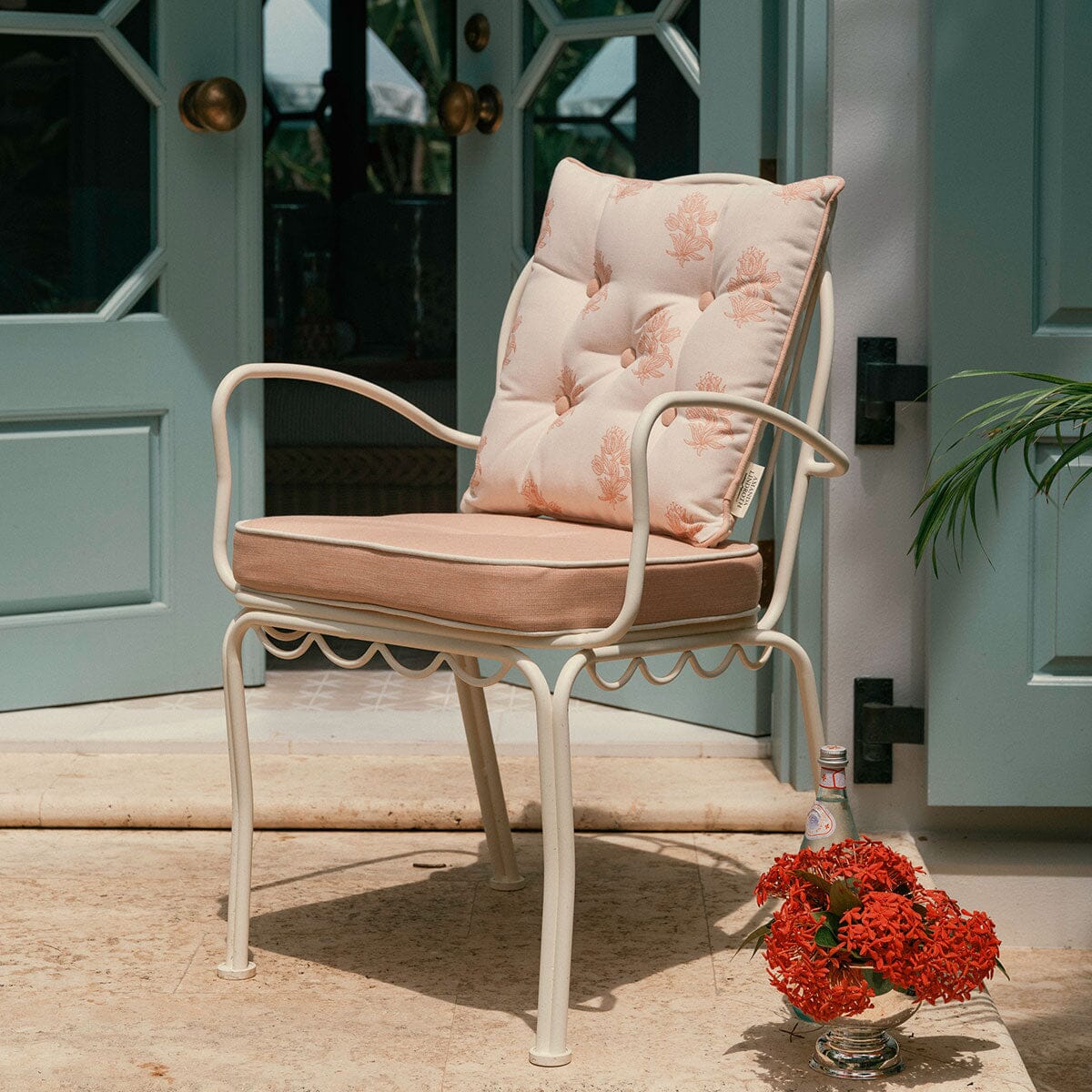 A white metal patio armchair with tufted cushions in a light beige and pink floral pattern, set on a stone patio in front of light blue doors.