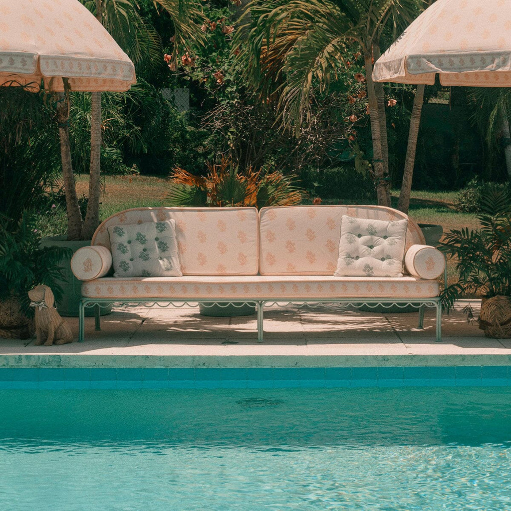 A cream and sage outdoor sofa with floral-patterned cushions sits beside a swimming pool, framed by tropical plants and two matching patio umbrellas.