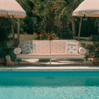 A cream and sage outdoor sofa with floral-patterned cushions sits beside a swimming pool, framed by tropical plants and two matching patio umbrellas.