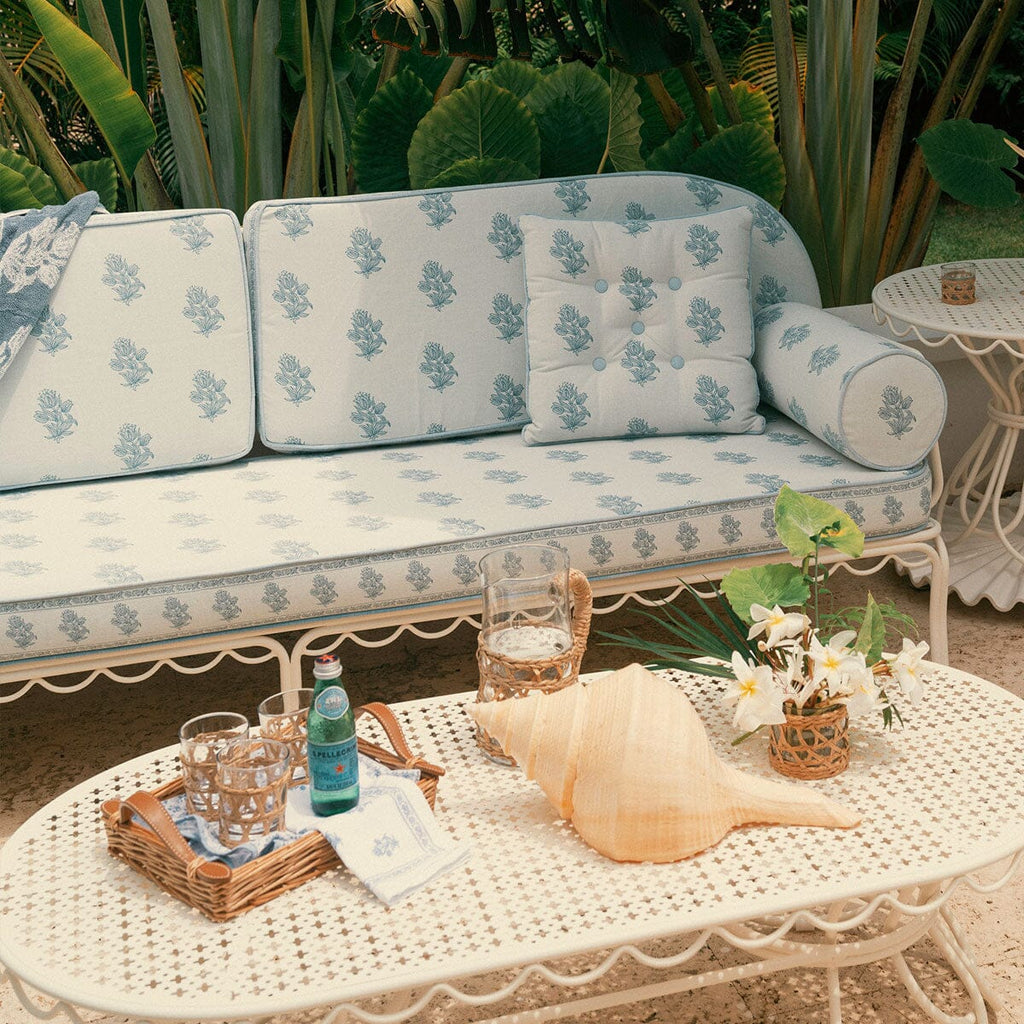 Outdoor patio furniture set with a white metal sofa and matching coffee table covered in light blue and white floral cushions, styled with a conch shell, glasses, and flowers in front of tropical green plants.