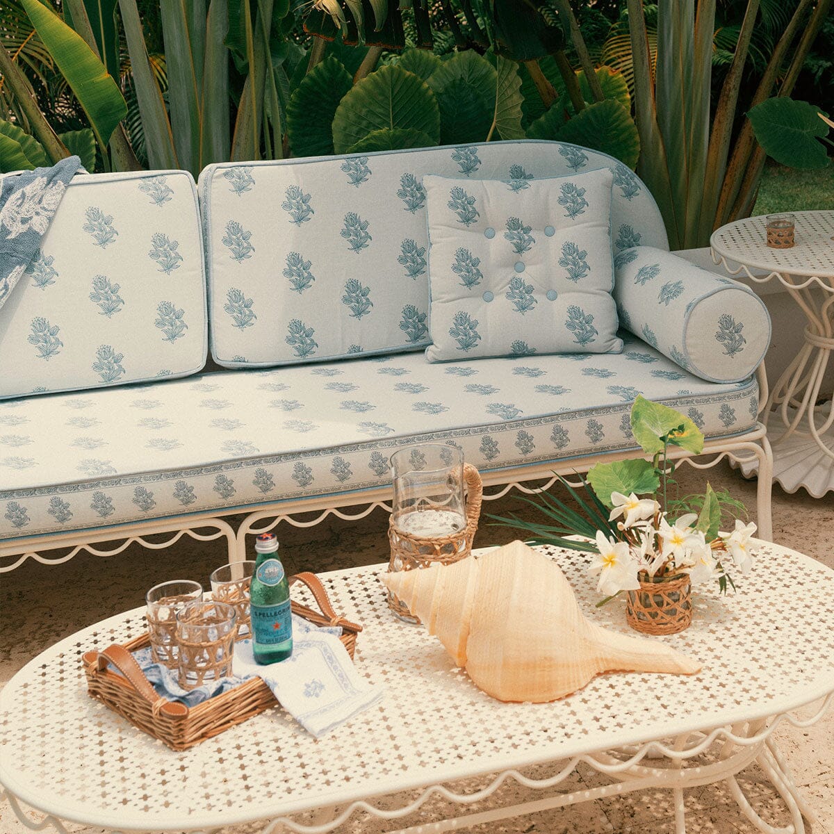 Outdoor patio furniture set with a white metal sofa and matching coffee table covered in light blue and white floral cushions, styled with a conch shell, glasses, and flowers in front of tropical green plants.