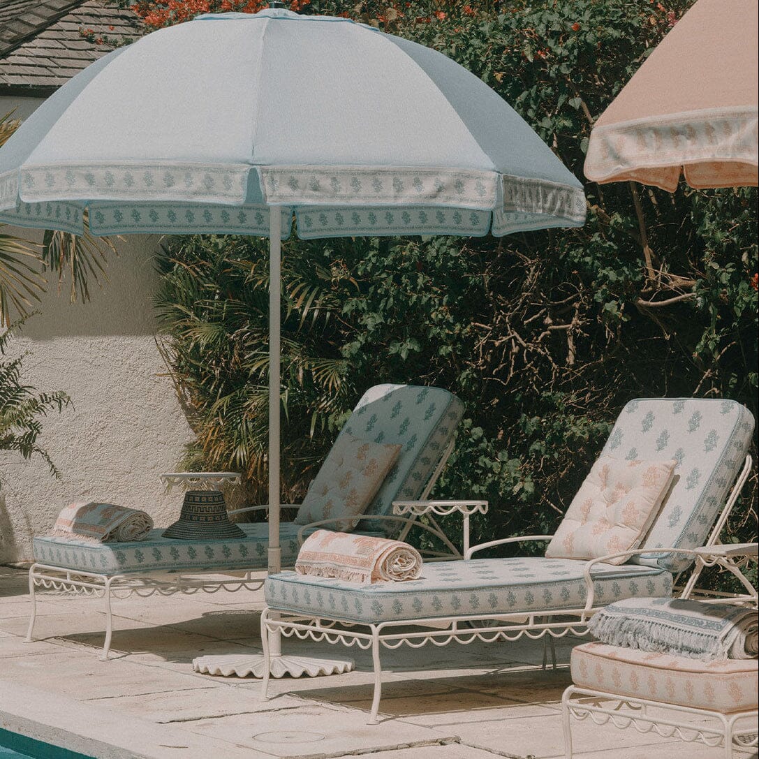 A light blue and white patterned outdoor patio umbrella shading two matching white lounge chairs with cream cushions, set beside a pool with tropical plants and a stucco wall in the background.