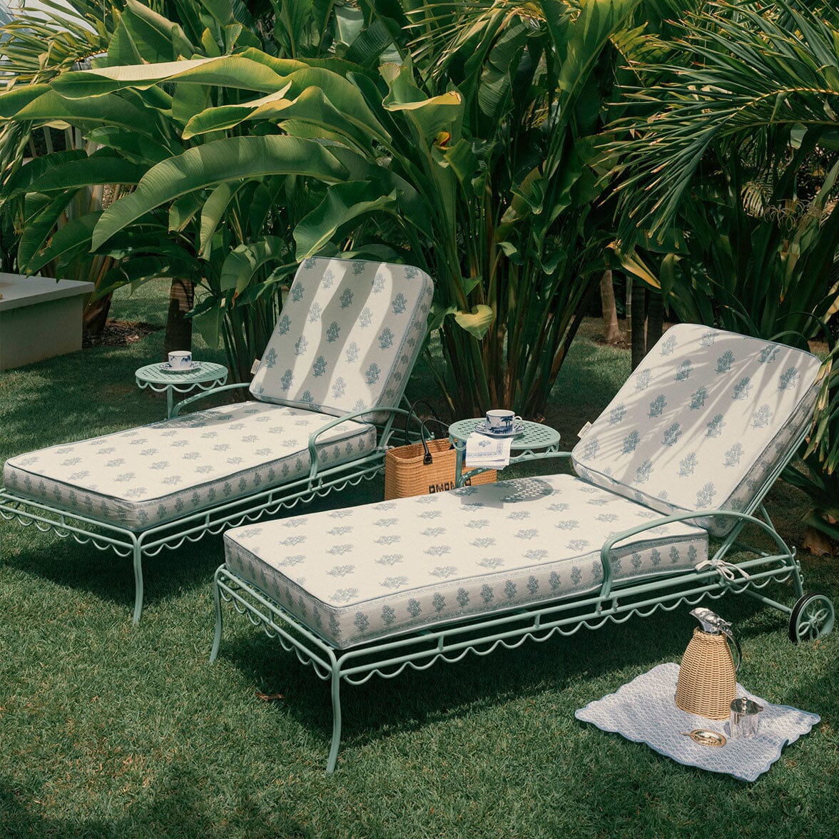 Two metal outdoor chaise lounge chairs with patterned cream cushions, set on a green lawn in front of large tropical plants. Small side tables sit between the chairs, and a woven bag and a small towel with a metal container are placed nearby.
