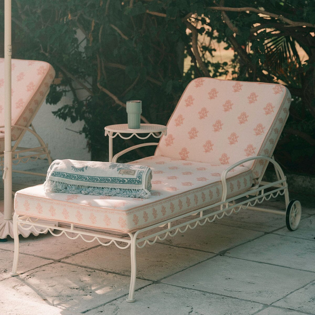 A white metal outdoor chaise lounge with pale pink patterned cushions, shown on a patio with a matching side table and folded blanket. Another similar lounge chair is partially visible on the left, with leafy greenery in the background.