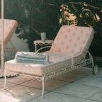 A white metal outdoor chaise lounge with pale pink patterned cushions, shown on a patio with a matching side table and folded blanket. Another similar lounge chair is partially visible on the left, with leafy greenery in the background.