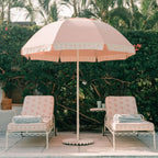 A pink patio umbrella with a scalloped white border stands between two matching pink lounge chairs with white metal frames on a patio, with a green hedge and palm trees in the background.