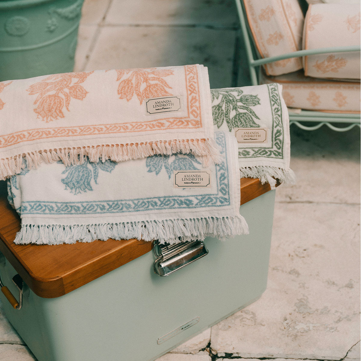 Three folded woven throw blankets with fringe, displayed on a mint green storage chest in a rustic setting. The blankets feature botanical leaf patterns in peach, light blue, and sage green on a cream background, with a small woven label reading 'Carmen's Handwoven Textiles'.