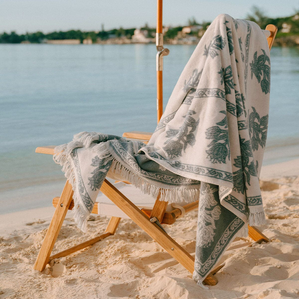 A patterned beach towel with fringed edges draped over a wooden lounge chair on a sandy beach, with calm water and distant trees in the background.