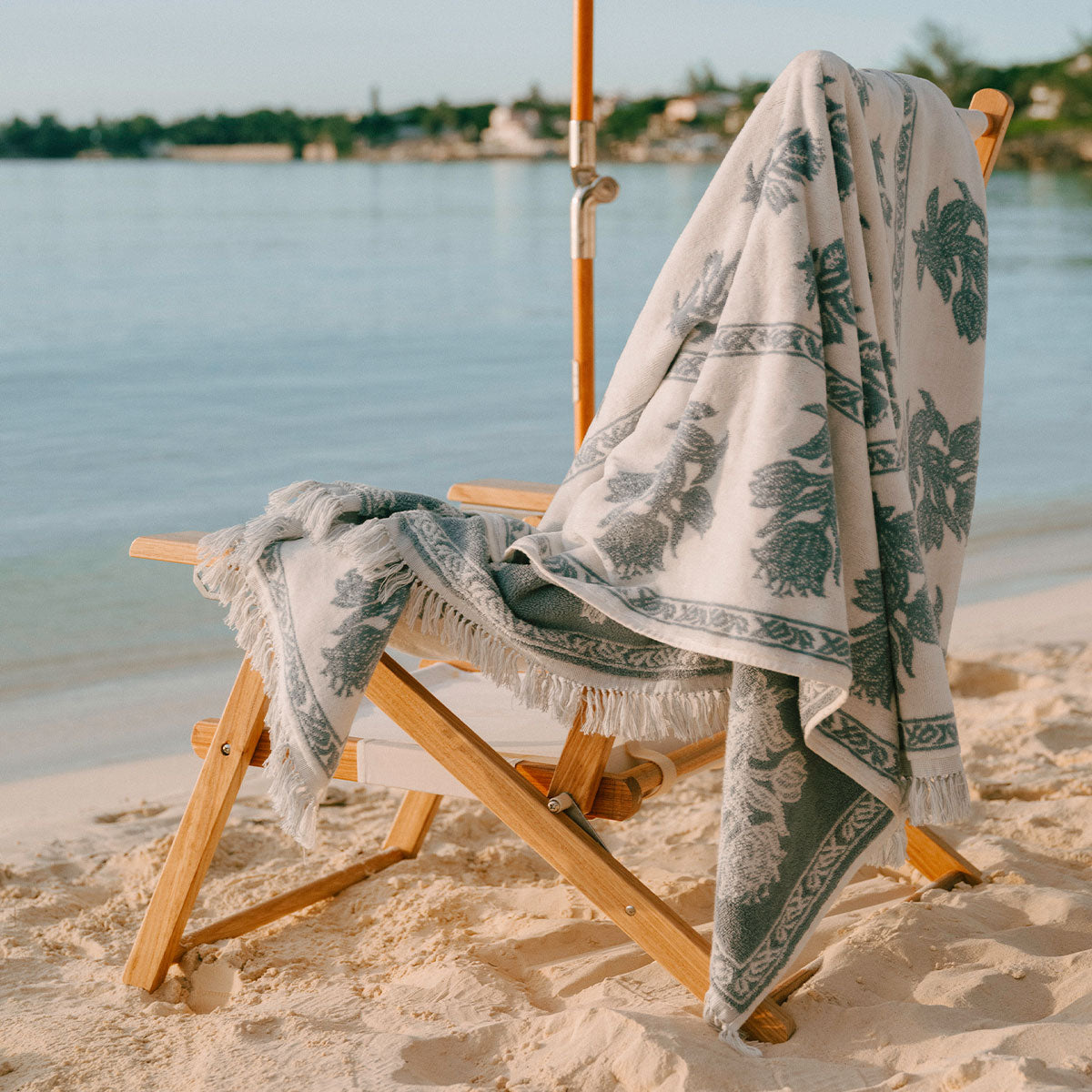 A patterned beach towel with fringed edges draped over a wooden lounge chair on a sandy beach, with calm water and distant trees in the background.