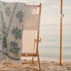 A white beach towel with a green botanical leaf pattern and fringed edge draped over a wooden beach chair on a sandy beach, with the ocean in the background.