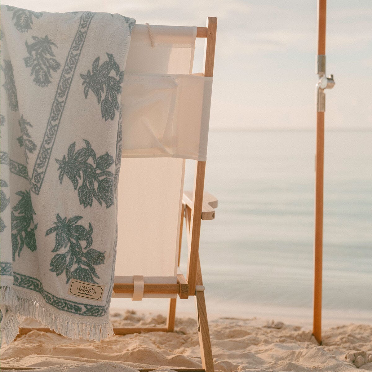 A white beach towel with a green botanical leaf pattern and fringed edge draped over a wooden beach chair on a sandy beach, with the ocean in the background.