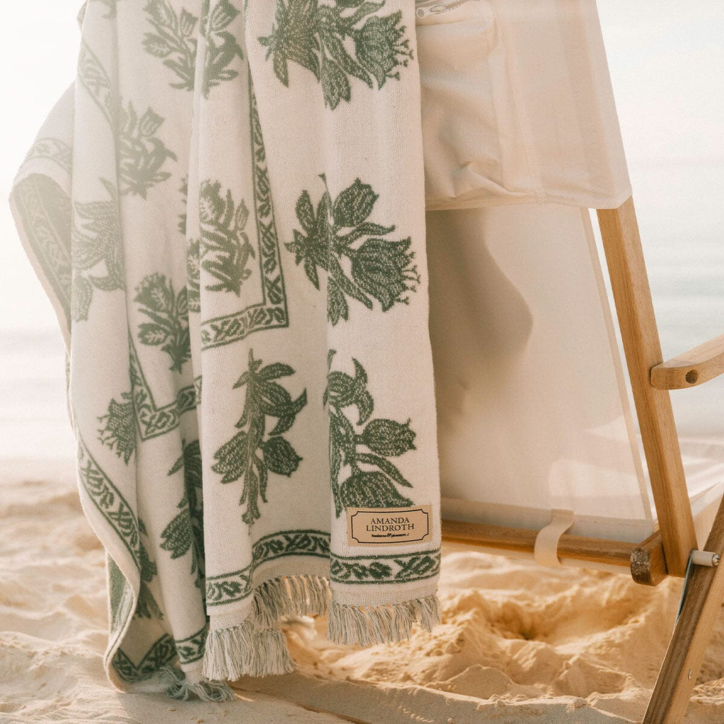 A white cotton beach towel with a green botanical leaf pattern and fringed edges, draped over a wooden beach chair on a sandy beach.