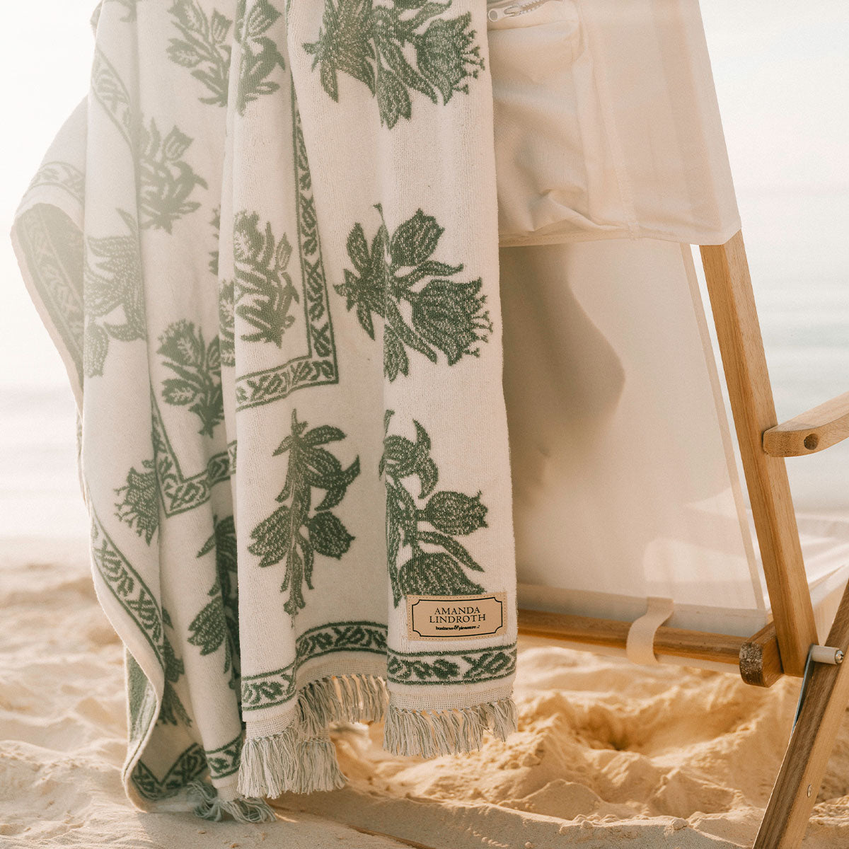 A white cotton beach towel with a green botanical leaf pattern and fringed edges, draped over a wooden beach chair on a sandy beach.