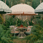 A styled outdoor patio scene with a central cream and peach patterned umbrella over a small round table and four matching cushioned chairs, surrounded by lush tropical greenery and partially visible side umbrellas.