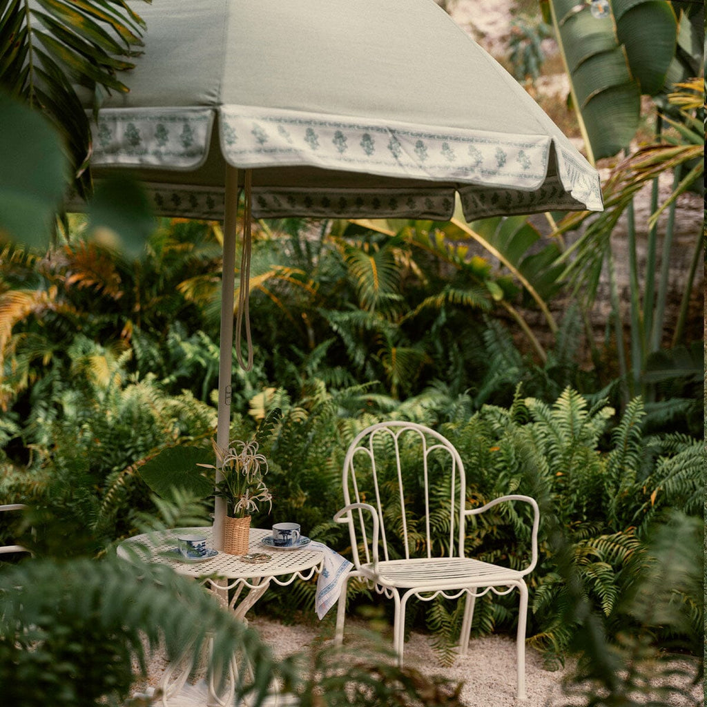 A white metal garden chair and matching small table sit beneath a large sage green patio umbrella with a floral patterned trim, surrounded by lush green tropical plants.