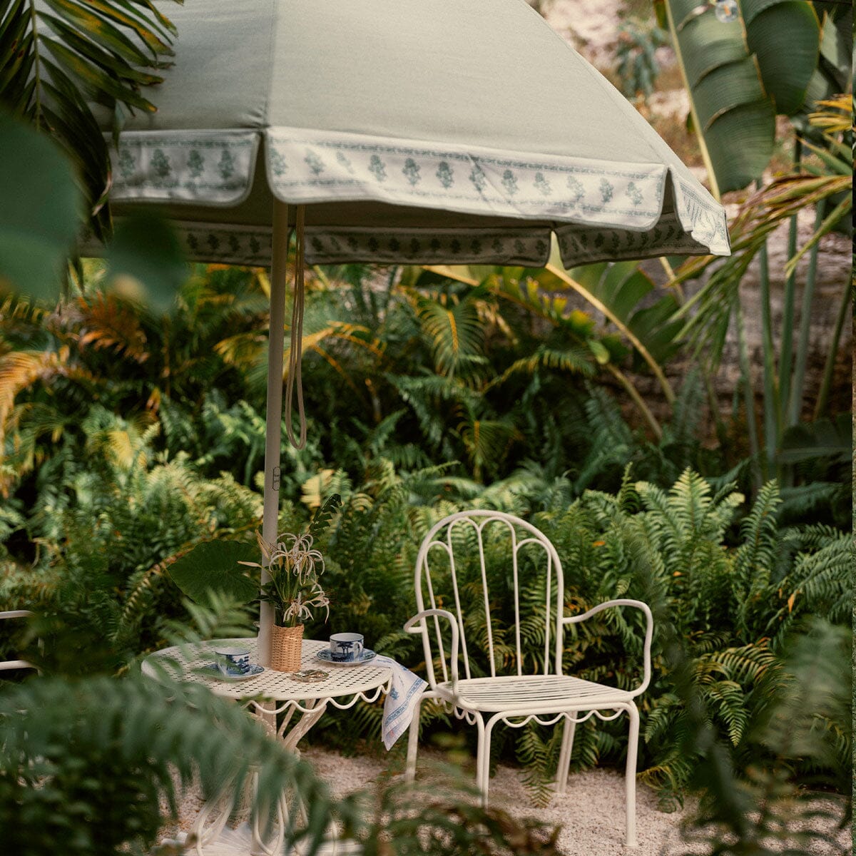 A white metal garden chair and matching small table sit beneath a large sage green patio umbrella with a floral patterned trim, surrounded by lush green tropical plants.