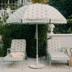 A patio scene with a large cream patio umbrella featuring a muted sage green floral pattern, flanked by two matching lounge chairs with a folded pink towel. The setting is outdoors on a stone patio with green plants and a white wall in the background.