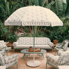 A coordinated outdoor patio furniture set with a large umbrella, sofa, chairs, and a round table, all in a matching light cream fabric with a pale green floral and border pattern, set against a lush tropical garden background.