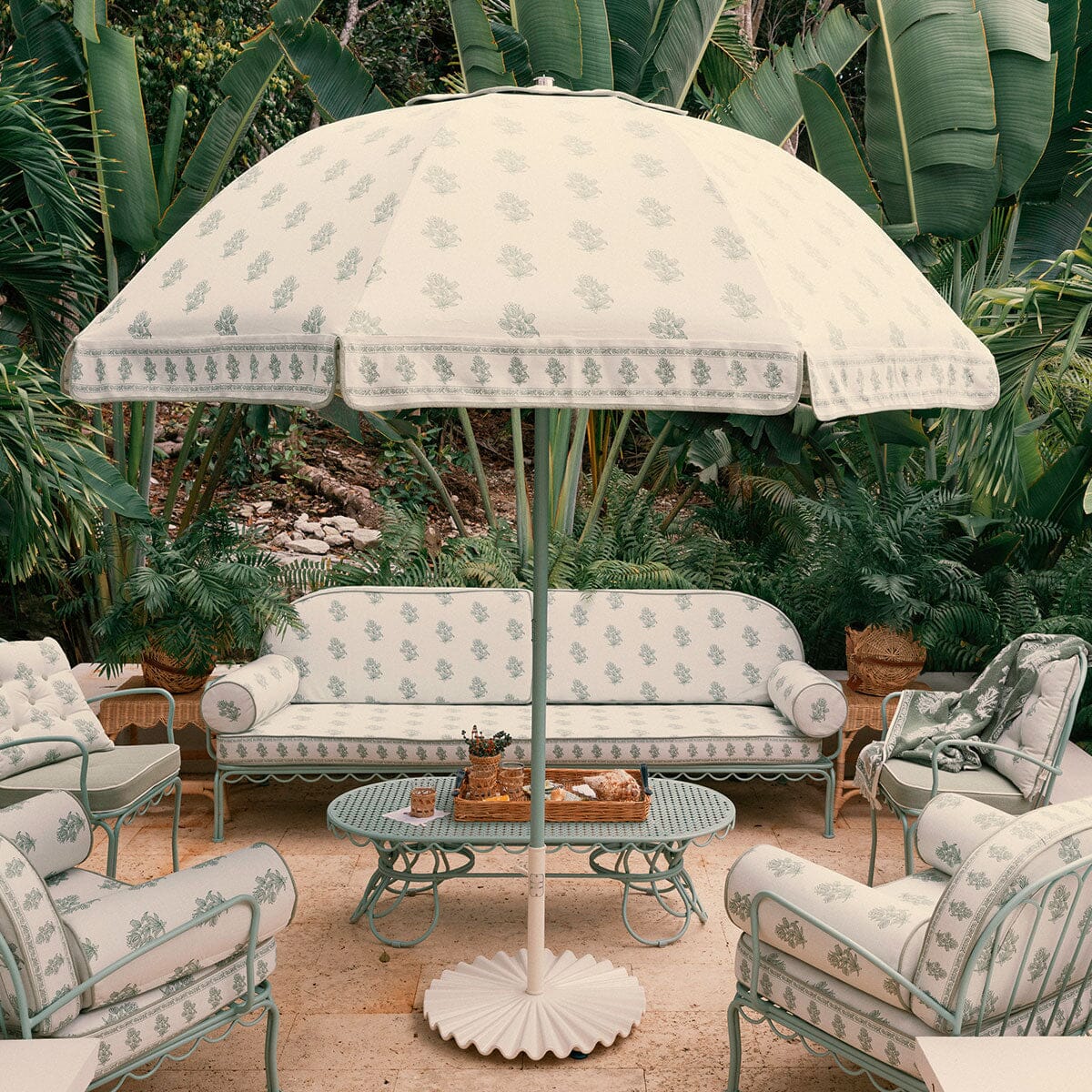 A coordinated outdoor patio furniture set with a large umbrella, sofa, chairs, and a round table, all in a matching light cream fabric with a pale green floral and border pattern, set against a lush tropical garden background.