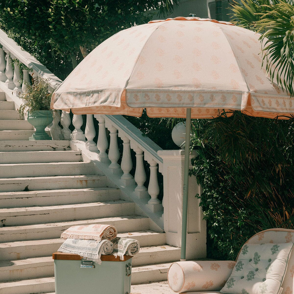 A large cream-colored patio umbrella with a delicate floral pattern and a matching cylindrical cushion, set on a sunlit outdoor staircase next to a white balustrade and lush green foliage.