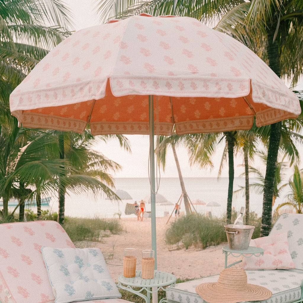 A beachside patio setup with a large white and coral sun umbrella, matching lounge chairs with patterned cushions, palm trees, and a sandy beach in the background.