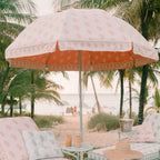 A beachside patio setup with a large white and coral sun umbrella, matching lounge chairs with patterned cushions, palm trees, and a sandy beach in the background.