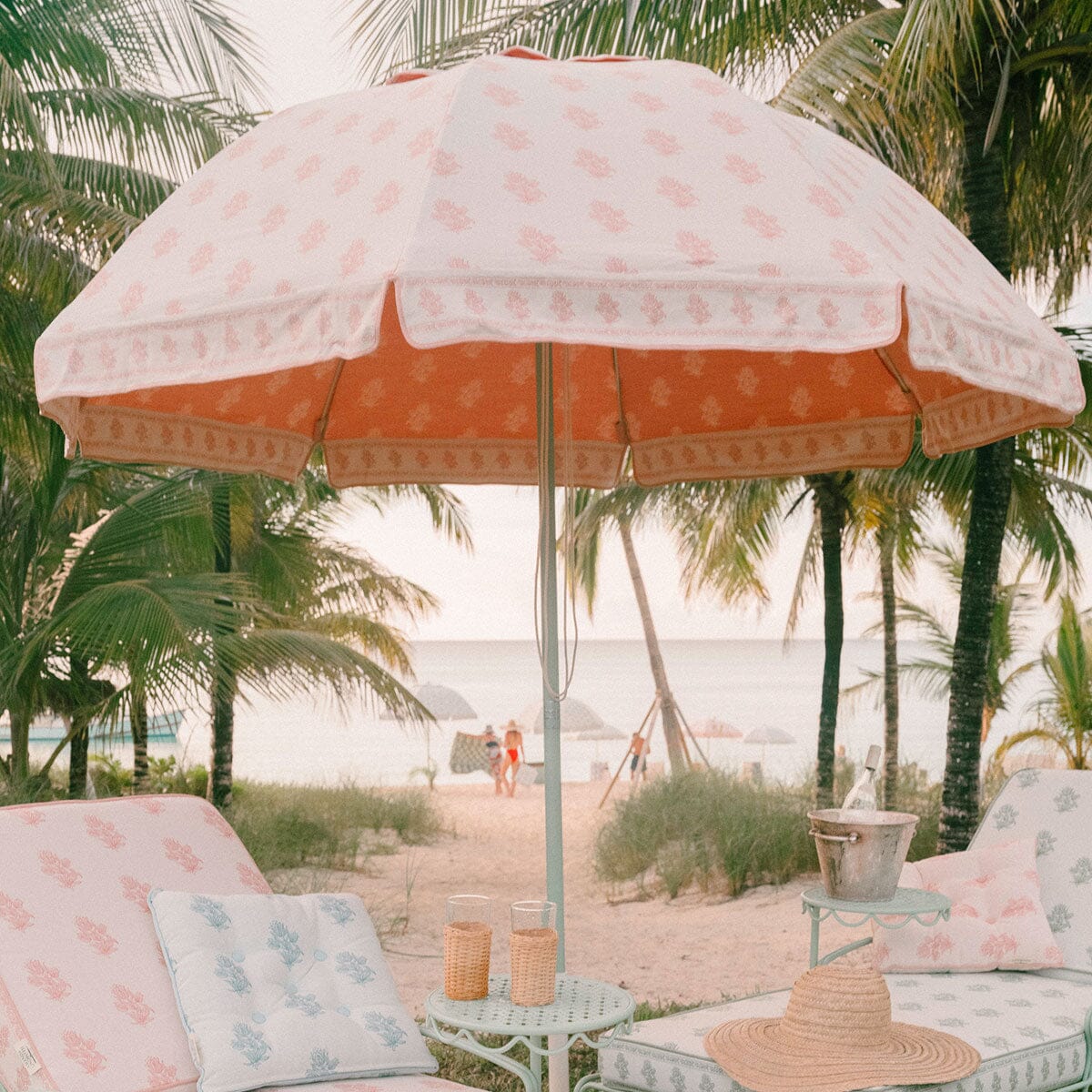A beachside patio setup with a large white and coral sun umbrella, matching lounge chairs with patterned cushions, palm trees, and a sandy beach in the background.