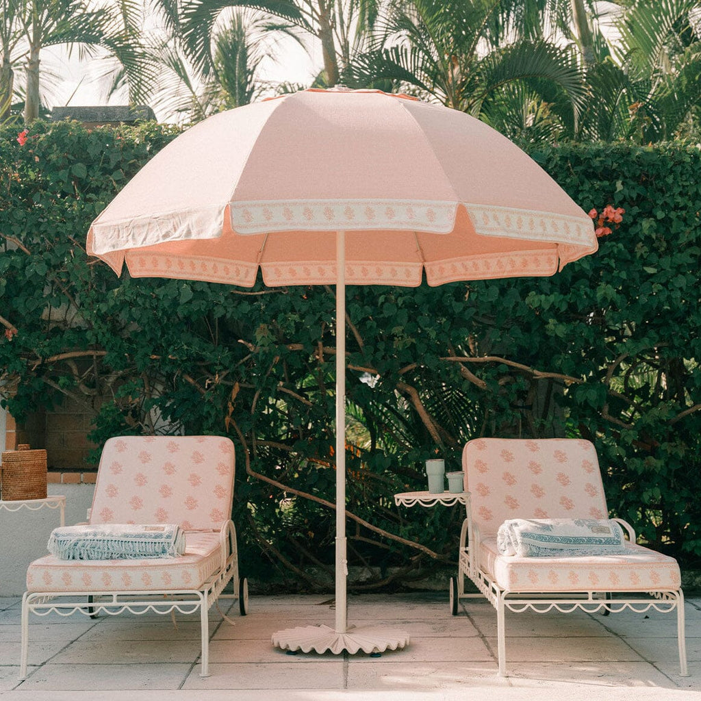A pink patio umbrella with a scalloped white border stands between two matching pink lounge chairs with white metal frames on a patio, with a green hedge and palm trees in the background.