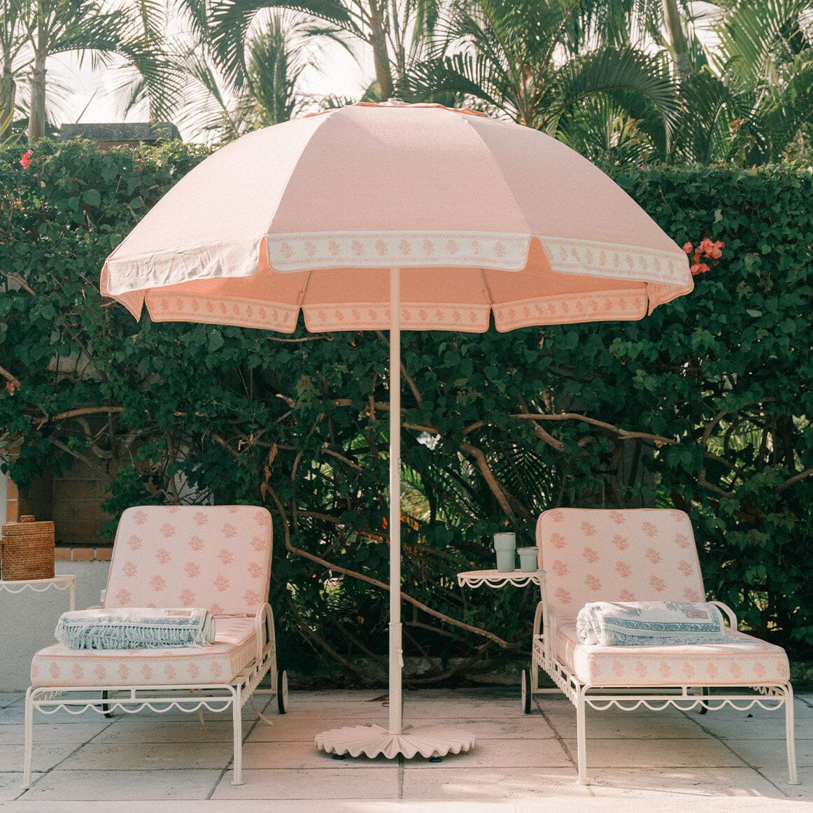 A pink patio umbrella with a scalloped white border stands between two matching pink lounge chairs with white metal frames on a patio, with a green hedge and palm trees in the background.